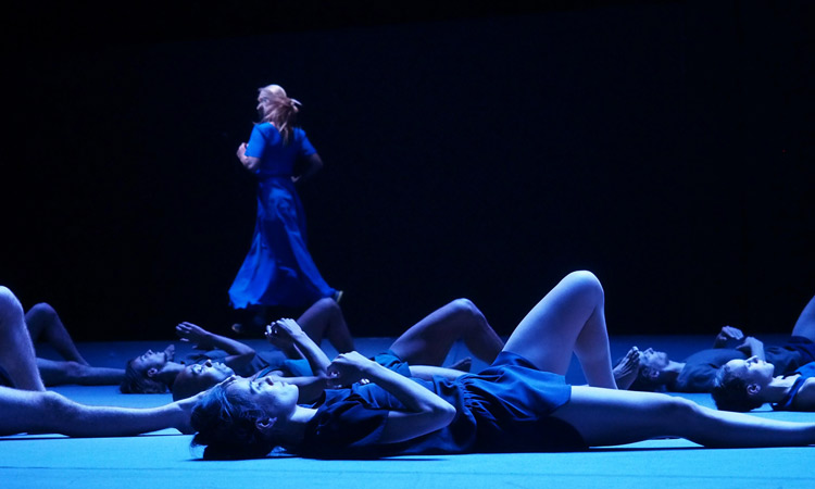 Dancers lay on the stage on their backs with their right legs extended straight on the floor and their left foot is on the ground so that their knee stands up to the ceiling. One dancer stands in the background.